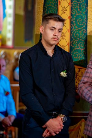 A person in a black suit jacket sits solemnly during what appears to be a formal ceremony.