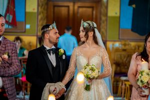 Couple participates in Orthodox wedding ceremony wearing ornate crowns in decorated church setting.