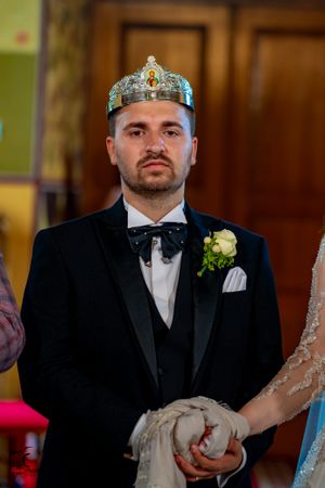 A groom wearing a crown and black tuxedo during an Orthodox Christian wedding ceremony.