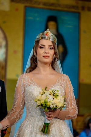 A bride in a beaded gown and crown holds a bouquet against a yellow and blue background.