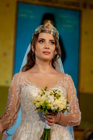 A bride wearing an ornate crown and sparkly dress holds a white floral bouquet.