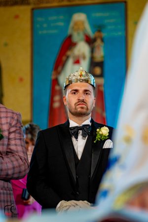 Groom wearing crown and black tuxedo during Orthodox wedding ceremony with religious artwork in background.