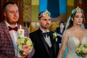 Wedding party during Orthodox ceremony with couple wearing traditional crowns and holding bouquets.
