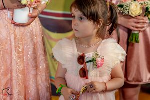 Young flower girl in pink dress with sunglasses and floral accessories at formal wedding.