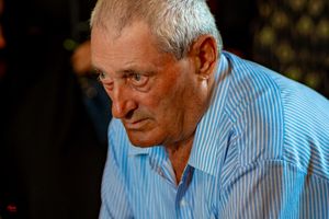An elderly person in a light blue striped shirt sits at a table during an event.