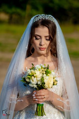 A bride in a lace veil holds a bouquet of yellow and white flowers in an outdoor setting.