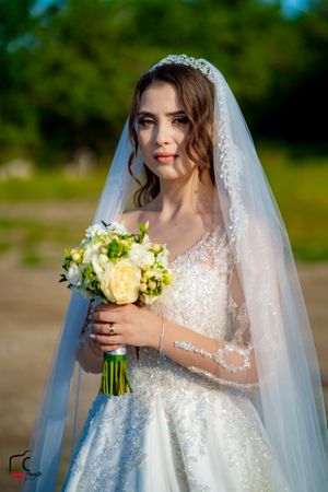 A bride in a flowing veil poses with her yellow bouquet against lush greenery.