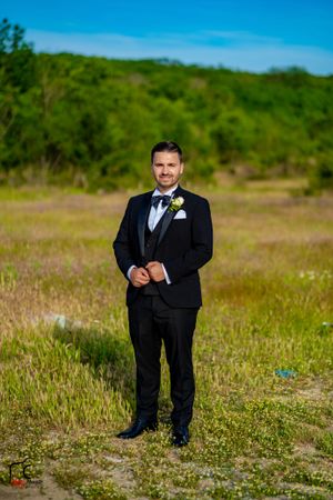 Groom in classic black tuxedo poses in a wildflower field under blue sky.