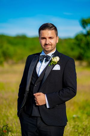 Groom wearing black tuxedo and bow tie stands in outdoor field setting.