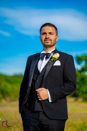 Formal groom portrait in black tuxedo with boutonniere against blue sky and green field.