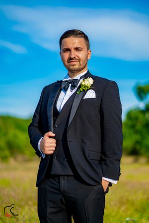 A groom in a classic black tuxedo poses outdoors against a bright blue sky and green field background.