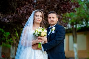 A romantic outdoor wedding portrait with a couple holding a bouquet of yellow flowers against a garden backdrop.