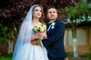 An elegant wedding couple poses together outdoors with a white bridal bouquet and formal attire.
