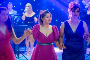 Three women in colorful dresses dancing together at a wedding reception under purple lights.