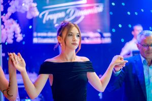 Elegant dancer in black off-shoulder dress performs at an event with blue stage lighting and neon signage.