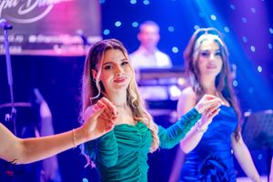 Performers in jewel-toned dresses dance in formation under dramatic blue and pink stage lighting.