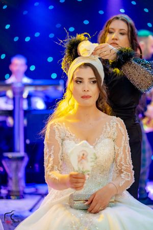 A stylist adjusts a bride's headpiece as she sits in an elegant white lace wedding gown.