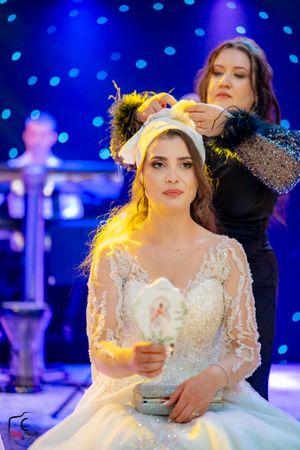 A bride in a detailed white gown sits patiently as her hair is styled under yellow lighting.