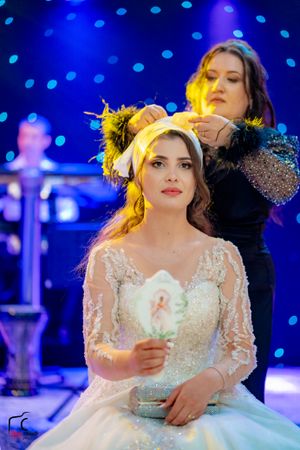A bride holds a candle while getting her hair styled under dramatic blue and yellow lighting.