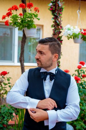 Groomsman in formal vest and bowtie adjusts his cuffs in a garden setting.
