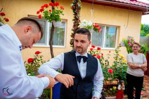 Wedding party member in formal attire stands near red flowers and yellow building exterior.