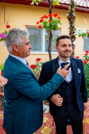 Two men in formal attire playfully interact in front of a yellow building with red flowers and greenery.