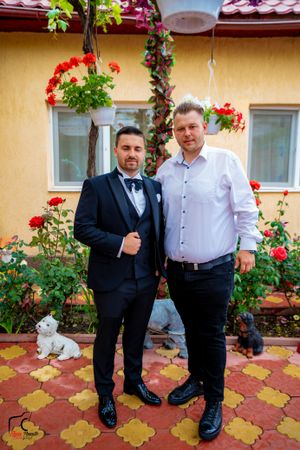 Two men standing on patterned tiles in front of yellow wall with hanging flower baskets.