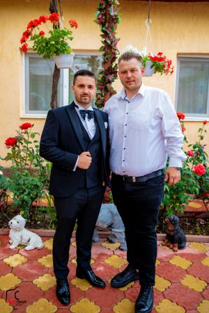 Two people in formal attire stand together on decorative tile flooring with red flowers and greenery in the background.