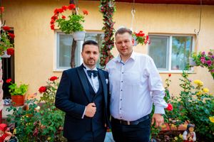 Two people pose together on a patterned patio with red geraniums and a beige wall behind them.