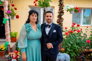 A couple in formal wear stands together against a yellow wall decorated with hanging flower baskets.