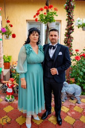 A couple poses together on a patio, with the person wearing a turquoise dress and another in a dark suit.