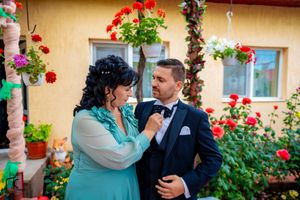 Two people in formal attire pose together in front of a garden wall with red flowering plants.