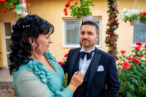 Two people in formal wear share a close moment against a backdrop of red flowers and greenery.