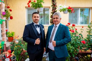 People in formal attire posing against yellow wall with hanging flower baskets.