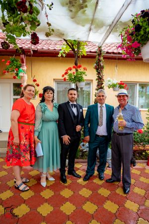Casual and formal dressed group posing on tiled patio with flowering plants and yellow walls.