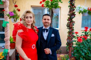 Couple posing in formal attire with red dress against floral garden backdrop.