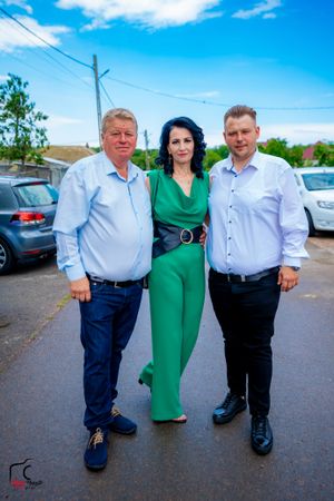 Three people in formal attire pose together outdoors in a parking lot on a sunny day.
