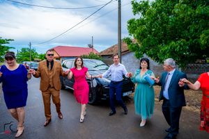 Party guests dance in a line on a street with cars and trees in the background.