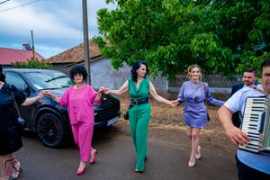 People in vibrant outfits dance in a line near parked cars and green trees.