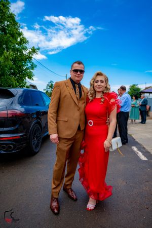 A couple dressed in brown suit and red dress pose together on a sunny day in a parking lot.