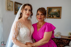 Bride in elegant lace gown shares special moment with wedding party member in bright room.