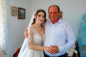A bride in an elegant lace gown poses with someone in formal attire against a light blue wall.