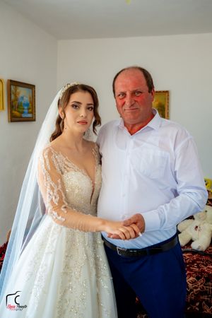 A bride in an elegant white lace dress stands with a companion in a white shirt and navy pants.