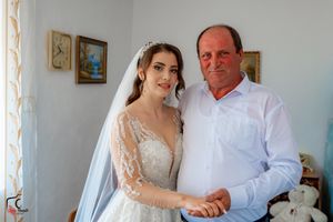 A bride in a lace wedding gown poses with a companion in a white dress shirt in a room with framed artwork.