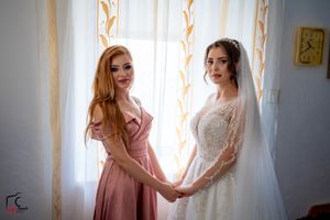 Two figures in formal attire holding hands by window with sheer curtains and natural lighting.