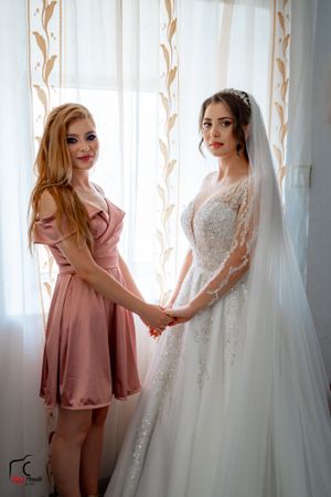 Two women admire an elegant white wedding dress hanging by a window with sheer curtains.