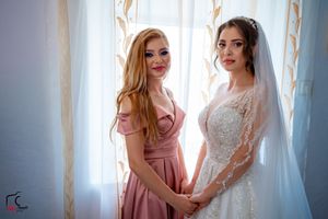 Two women in pink and white dresses examine wedding gown details near a sunlit window.