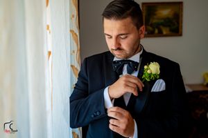Groom preparing for wedding in formal black tuxedo near window light.