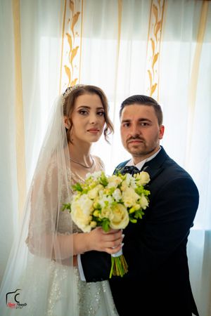 A newlywed couple poses together with a yellow and white floral bouquet against sheer curtains.