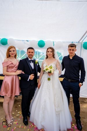 Wedding party members in formal attire gather under a white tent with teal decorations.
